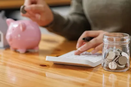 Pessoa a colocar moeda num mealheiro, junto a um frasco de poupanças e uma casa em miniatura